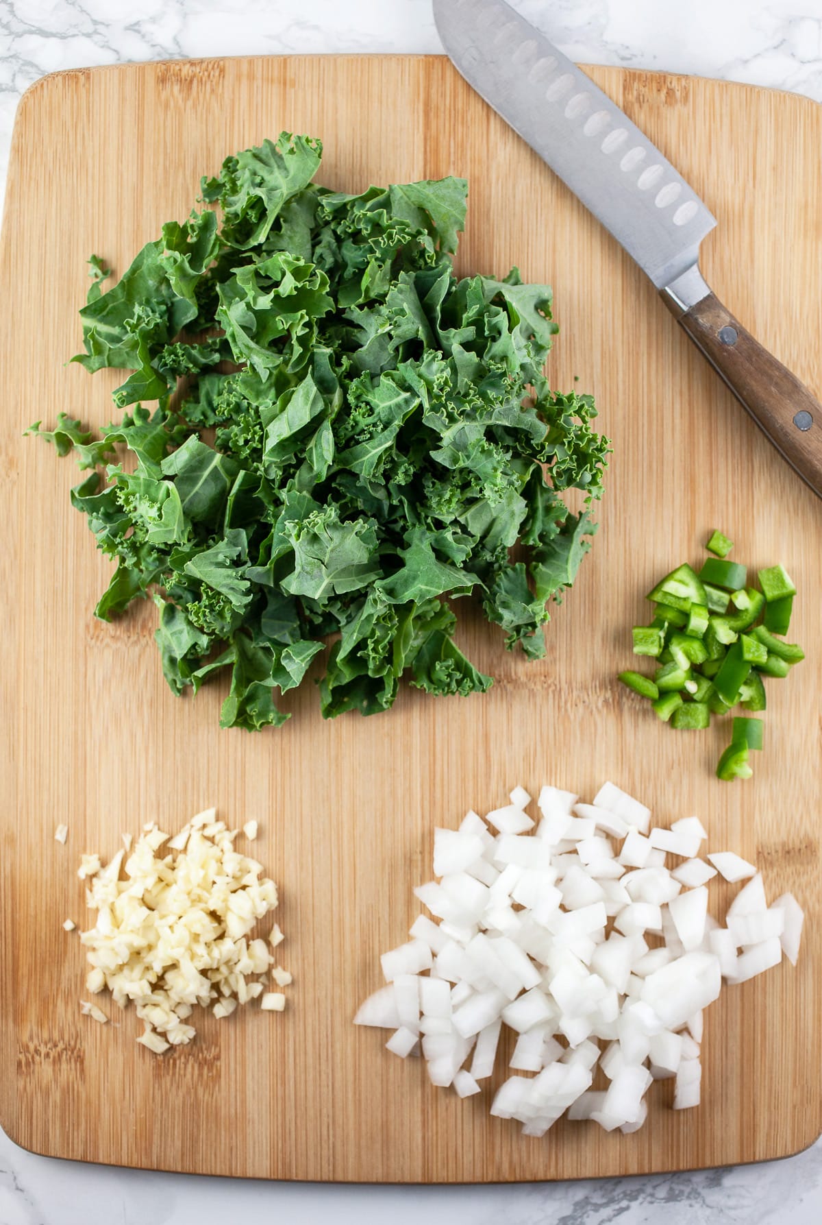 Minced garlic, onion, and jalapeno pepper and chopped kale on wooden cutting board with knife.