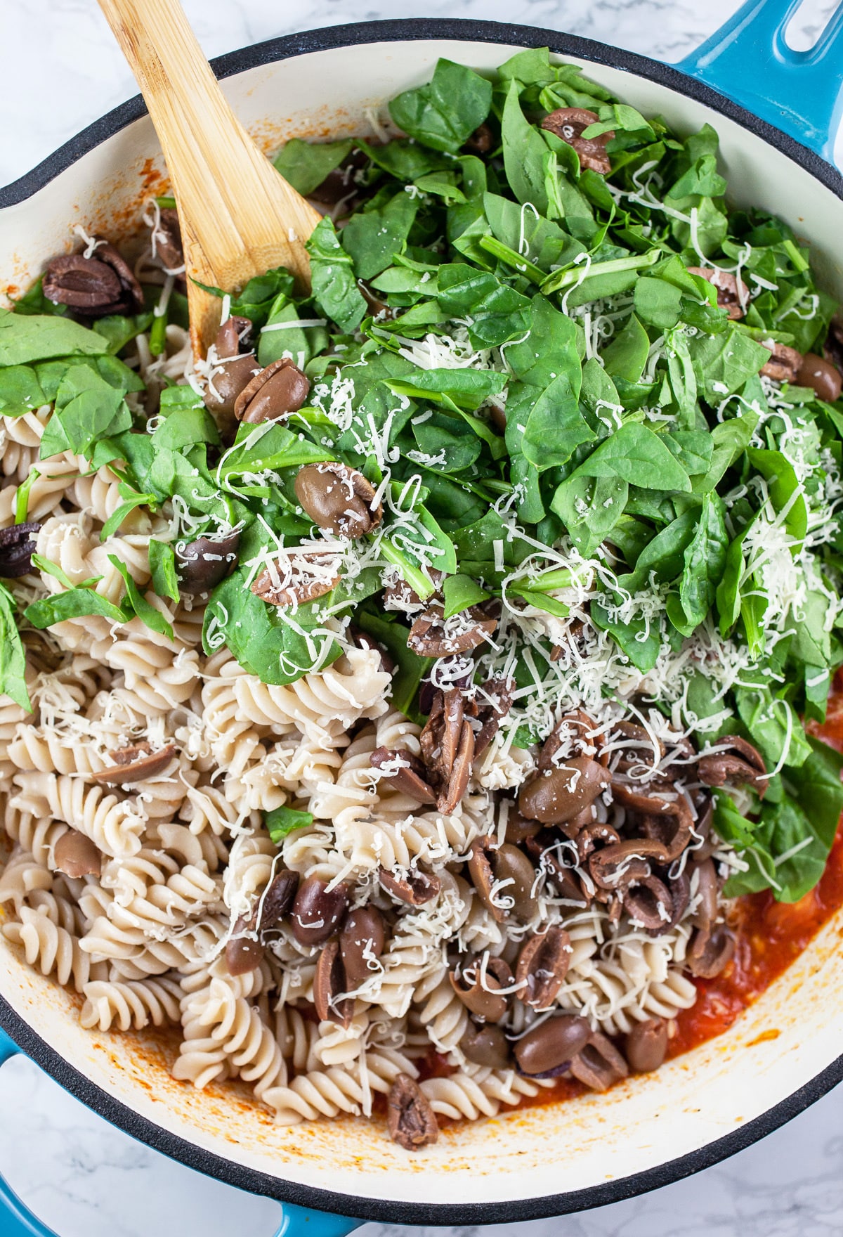 Cooked pasta, spinach, Kalamata olives, tomatoes, and Parmesan cheese in skillet with wooden spoon.