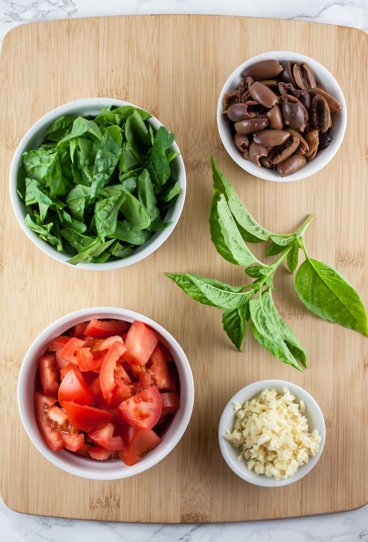 Diced tomatoes, spinach, Kalamata olives, garlic, and basil on wooden cutting board.