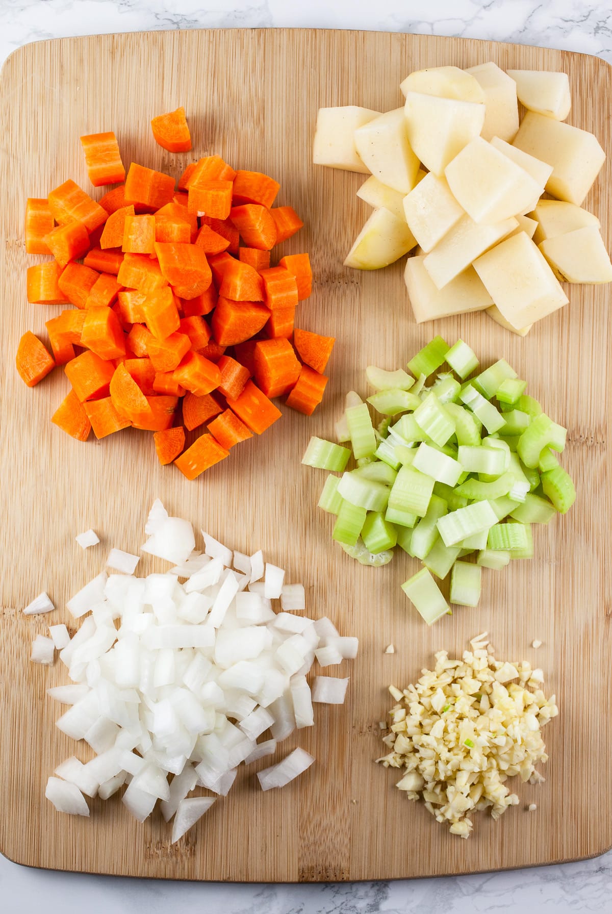 Minced garlic, onions, celery, and chopped carrots and potatoes on wooden cutting board.