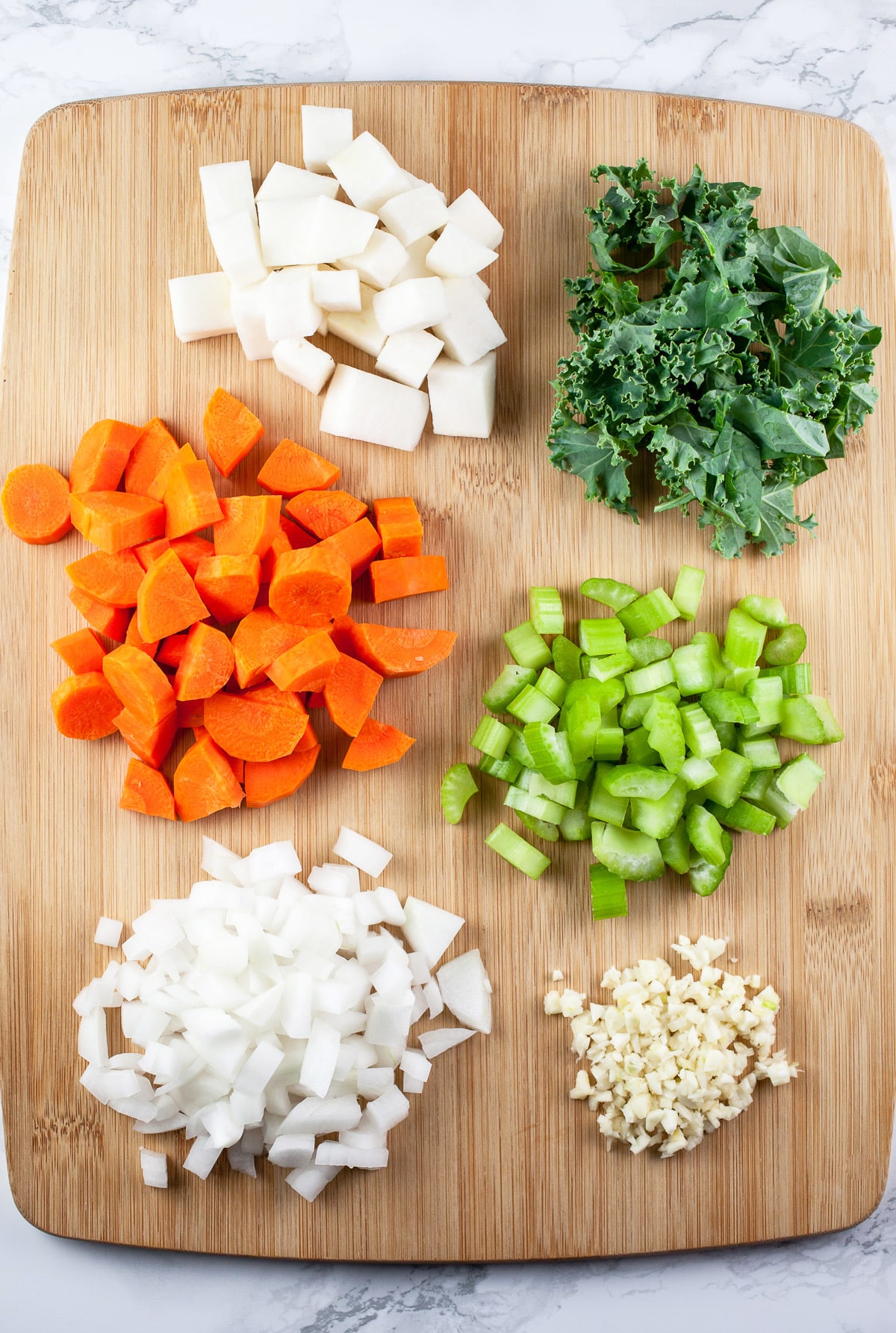 Minced garlic and onions, chopped carrots, celery, turnips, and kale on wooden cutting board.