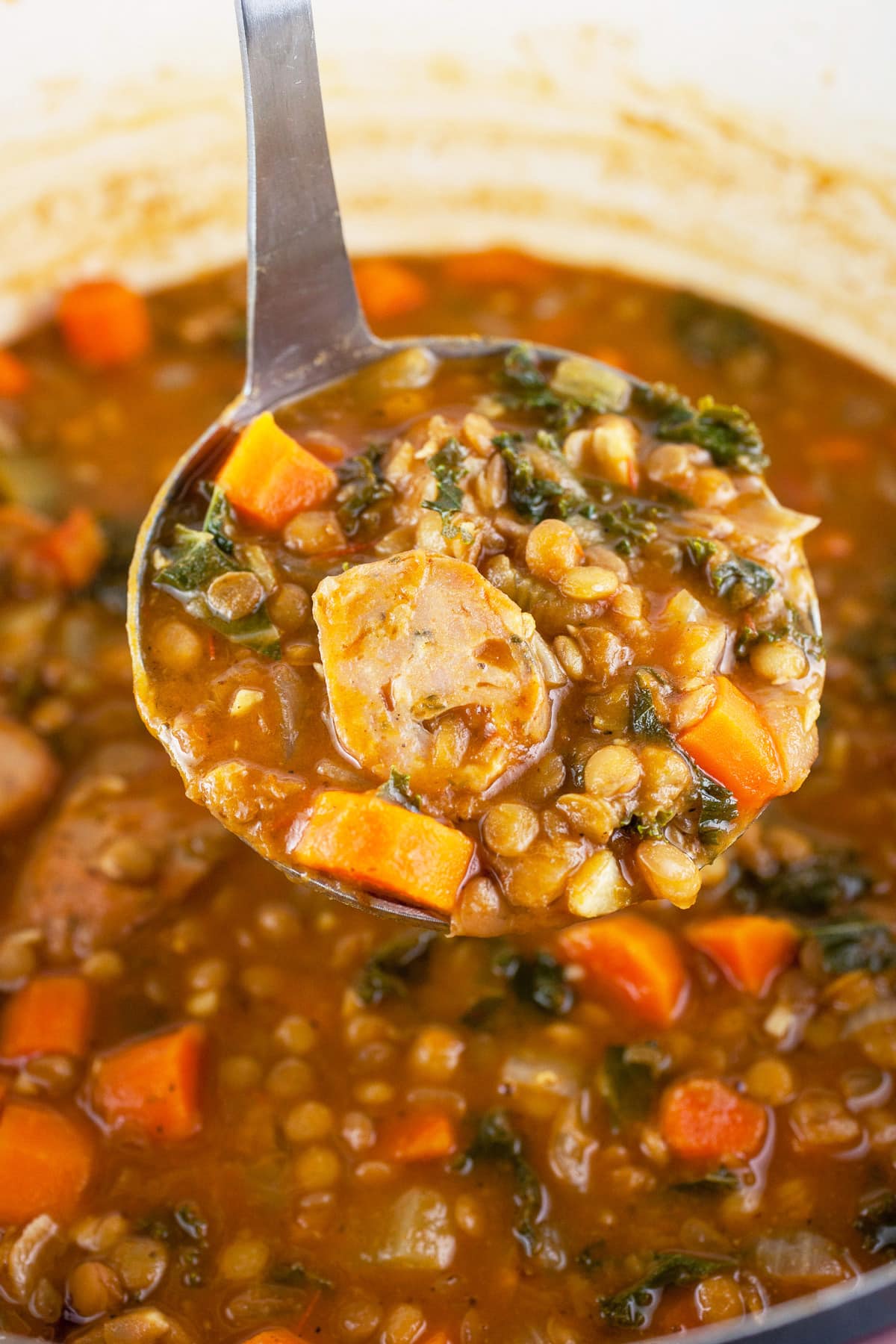 Scoop of sausage lentil soup lifted from Dutch oven on metal ladle.