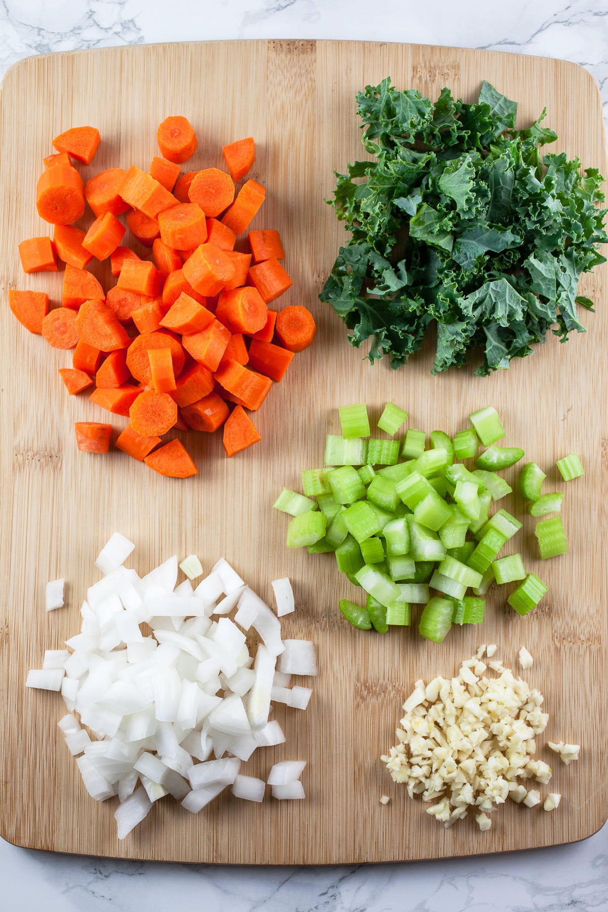 Minced garlic, onions, celery, carrots, and chopped kale on wooden cutting board.