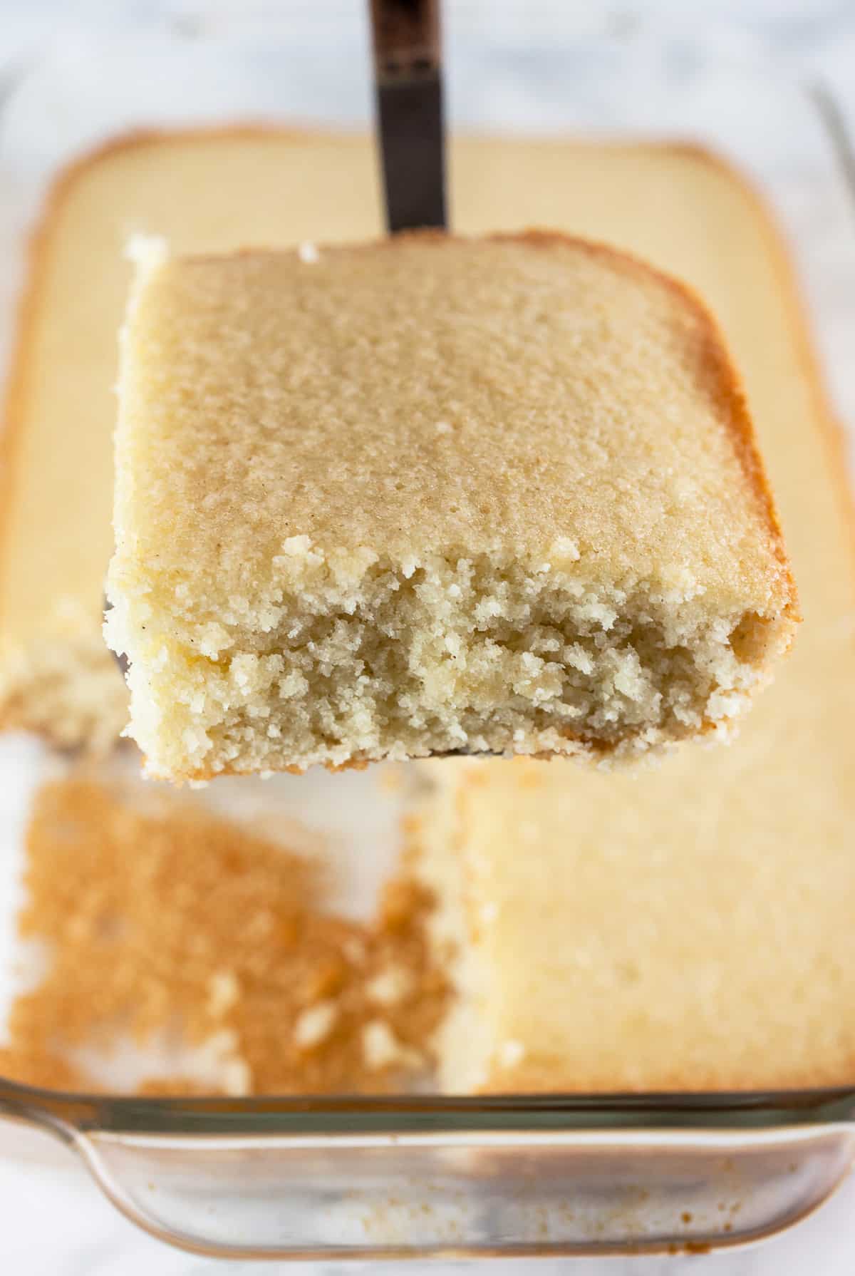 Slice of almond cake lifted from glass cake pan on metal spatula.