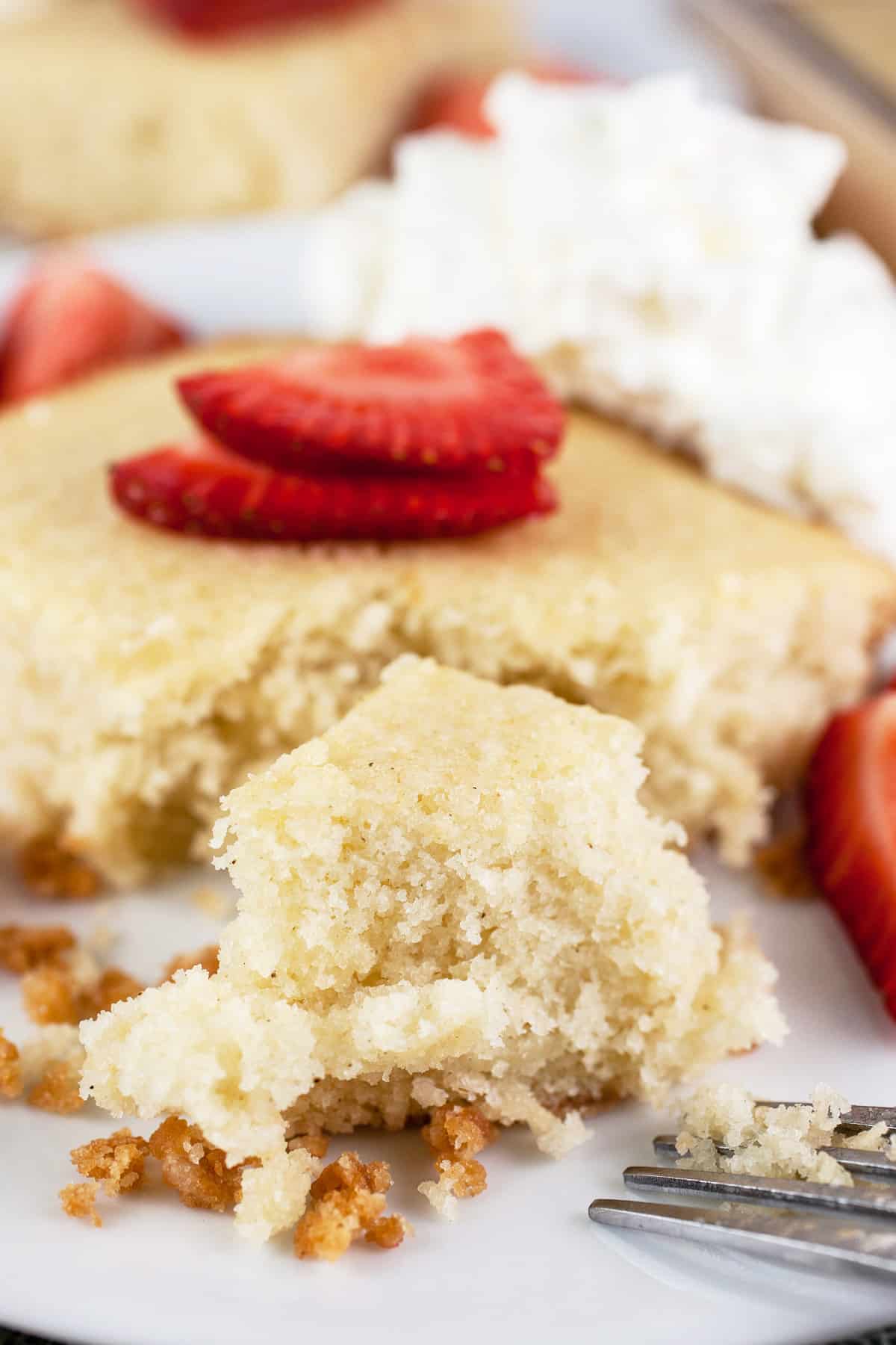 Bite cut from slice of almond cake on white plate with whipped cream and strawberries.