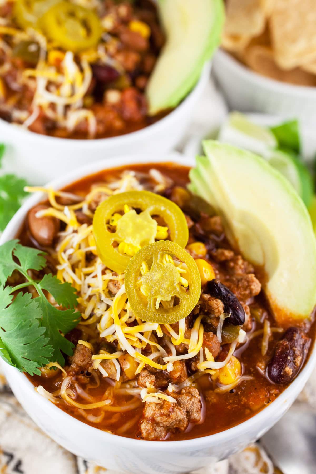Slow cooker turkey chili in white bowls with sliced avocado, cilantro, shredded cheese, and pickled jalapeno peppers.