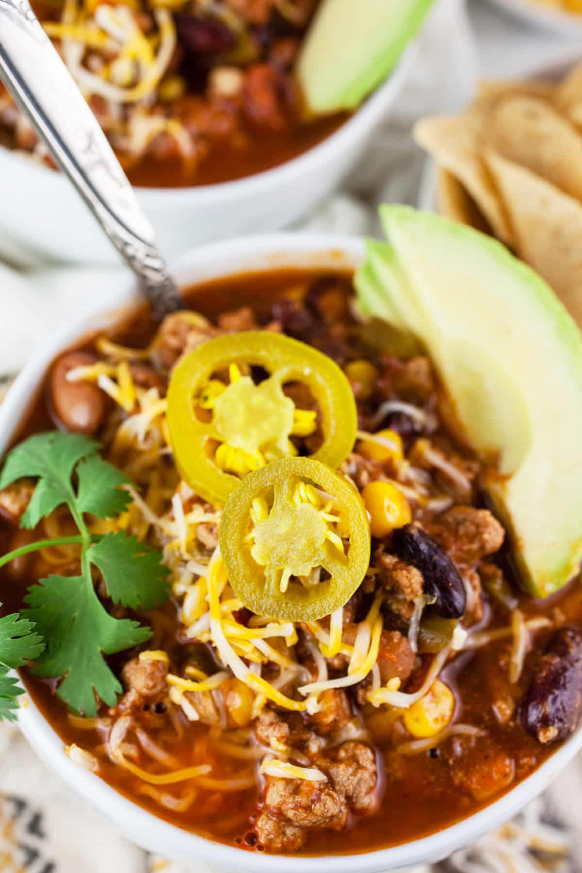 Turkey chili in white bowls with sliced avocado, cilantro, shredded cheese, and pickled jalapenos.