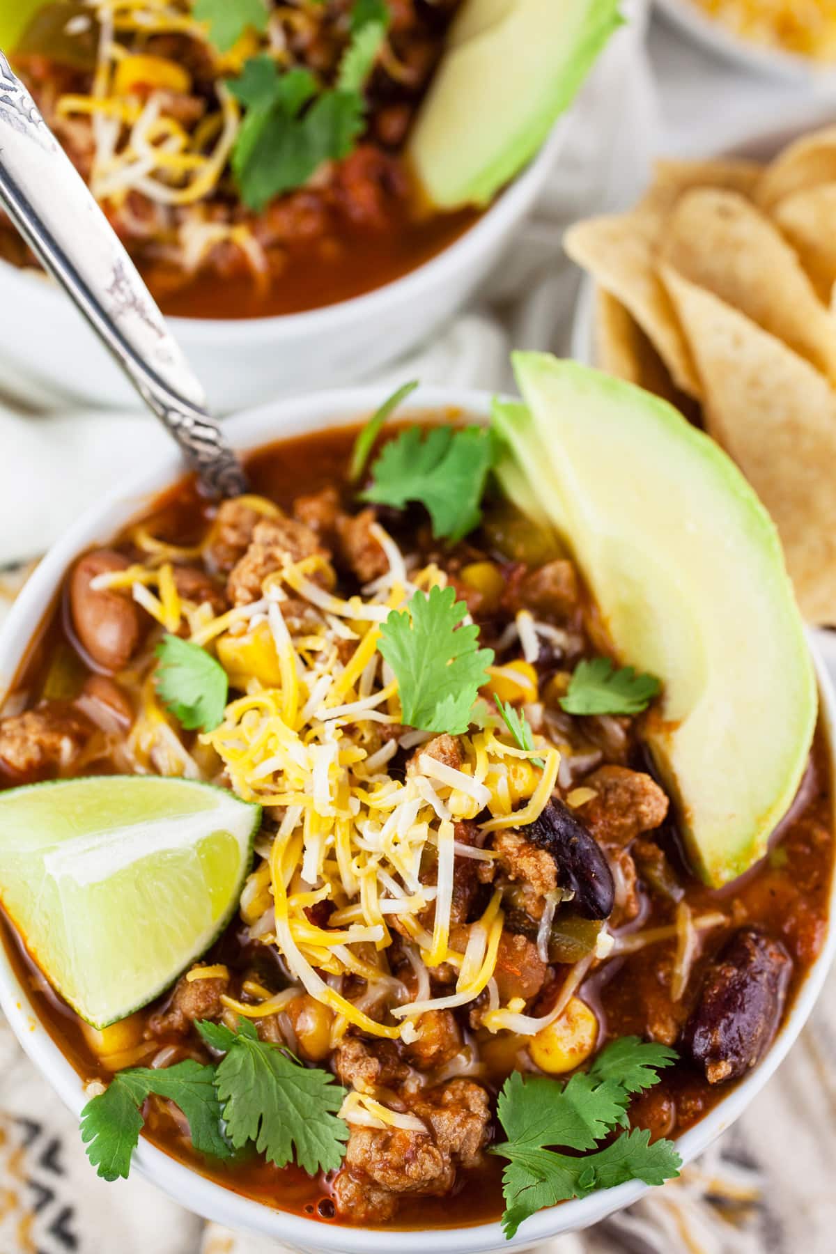 Slow cooker turkey chili in white bowls with sliced avocadoes, lime wedge, shredded cheese, and minced cilantro.