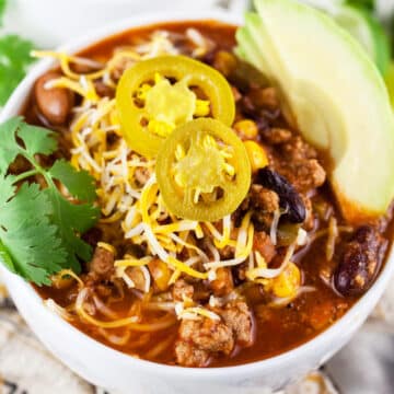 Bowl of slow cooker turkey chili with sliced avocado, shredded cheese, cilantro, and pickled jalapenos.