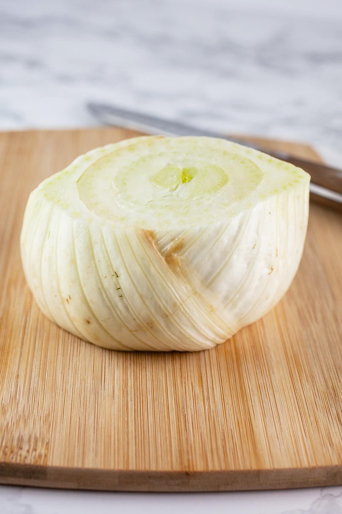 Top and bottom cut off from bulb of fennel on wooden cutting board with knife.