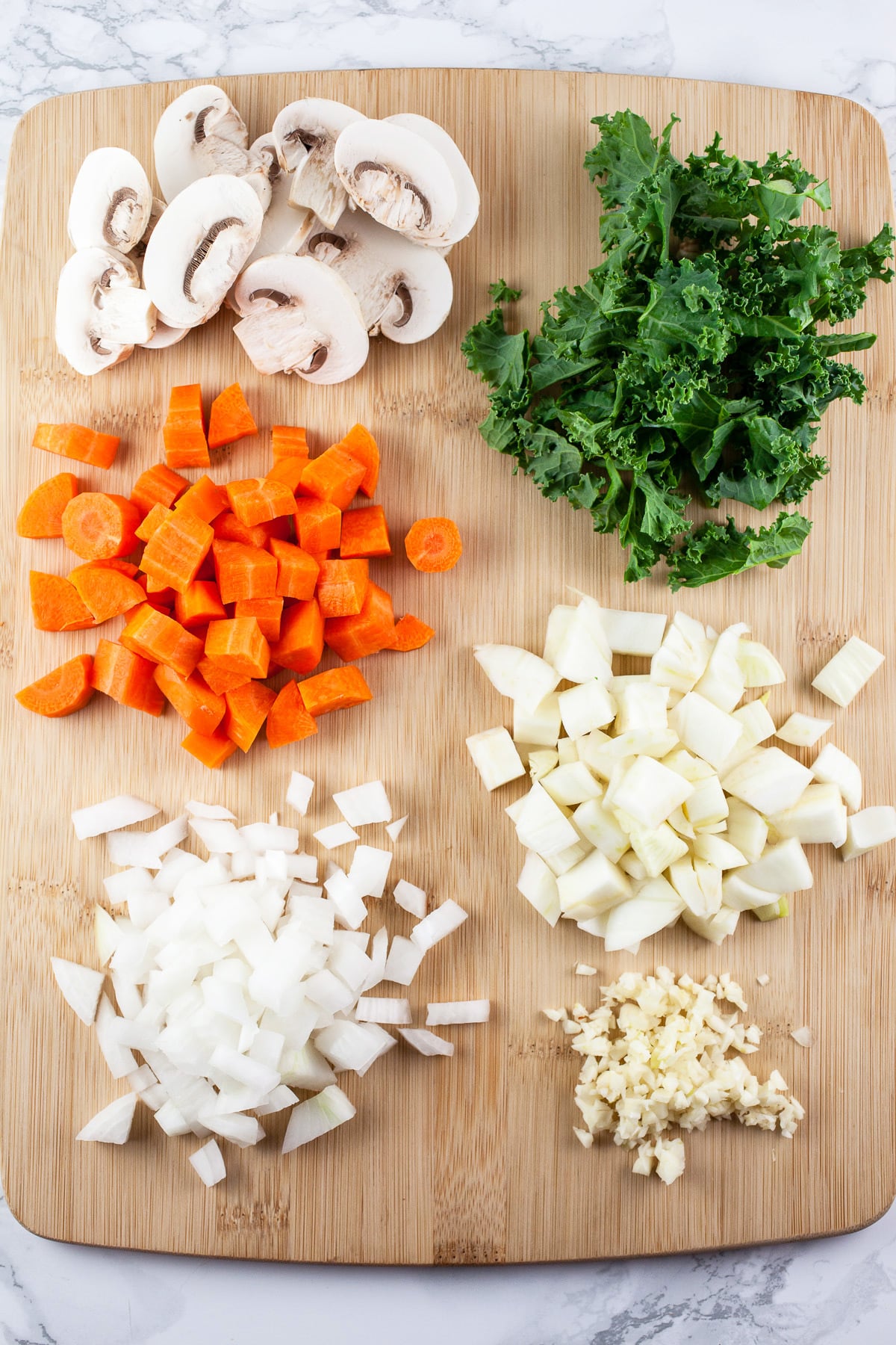 Minced garlic, onions, fennel, carrots, mushrooms, and kale on wooden cutting board.