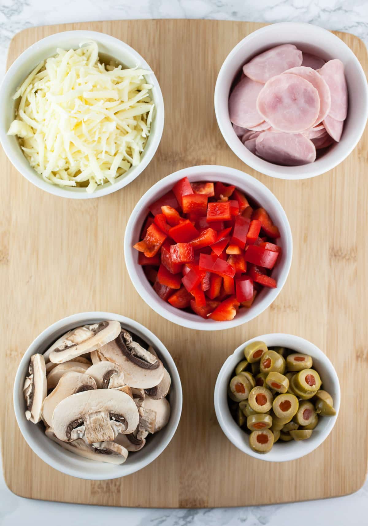 Sliced mushrooms, green olives, red bell peppers, Canadian bacon, and shredded cheese in white bowls on wooden cutting board.