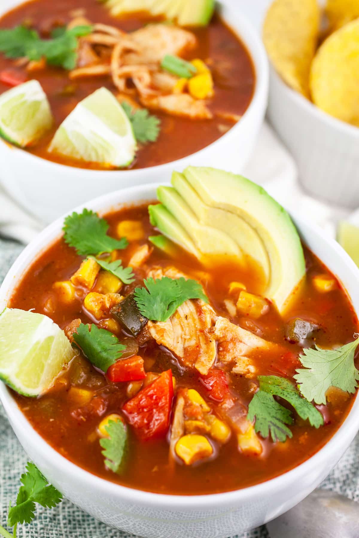 Chicken corn soup in white bowls with sliced avocado, cilantro, and lime wedges.