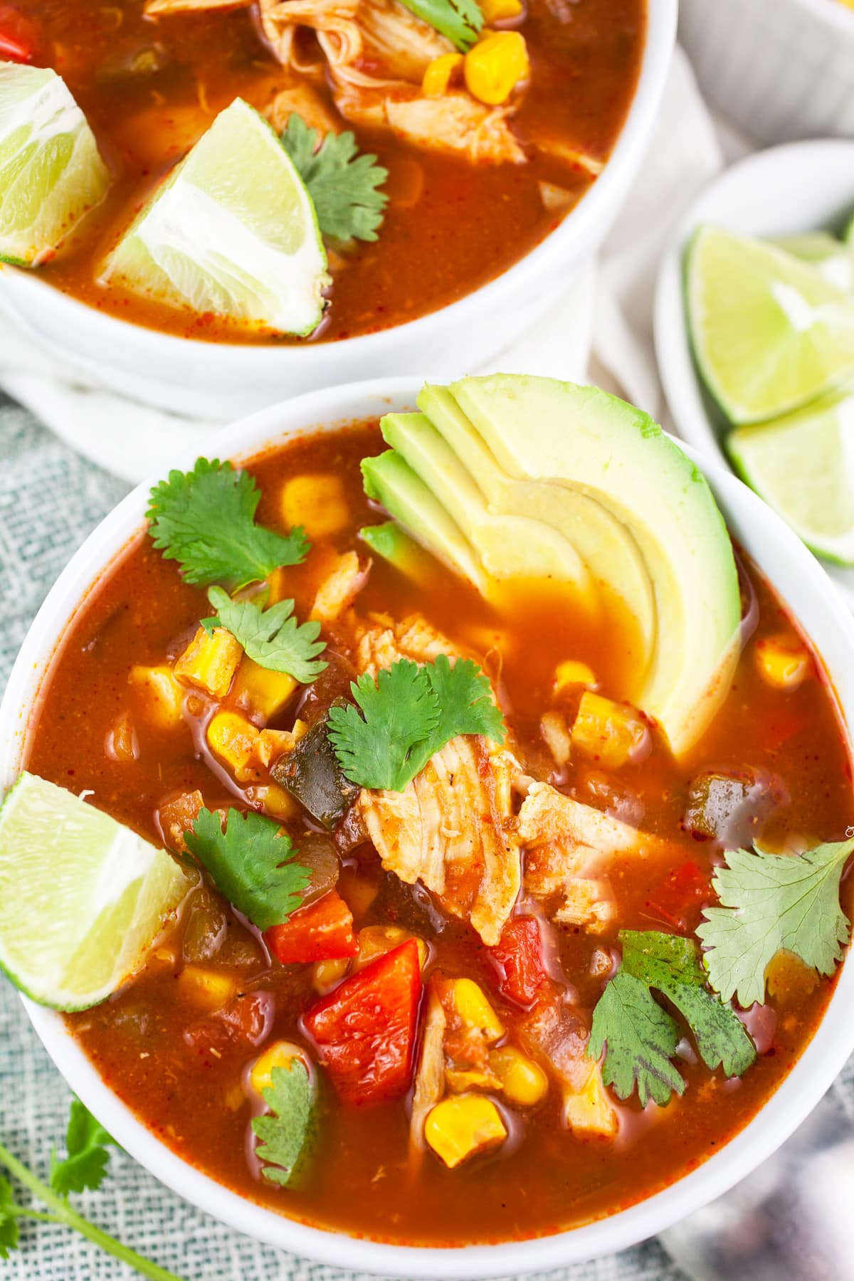 Southwest chicken corn soup in white bowls with sliced avocado, cilantro, and lime wedges.