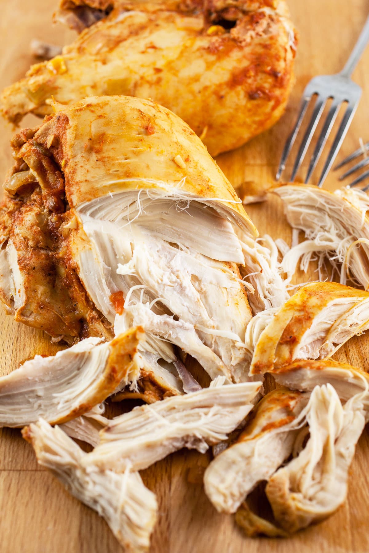 Cooked chicken breasts shredded with two forks on wooden cutting board.