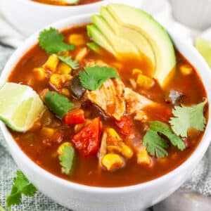 Chicken corn soup in white bowls with sliced avocado, cilantro, and lime wedge.