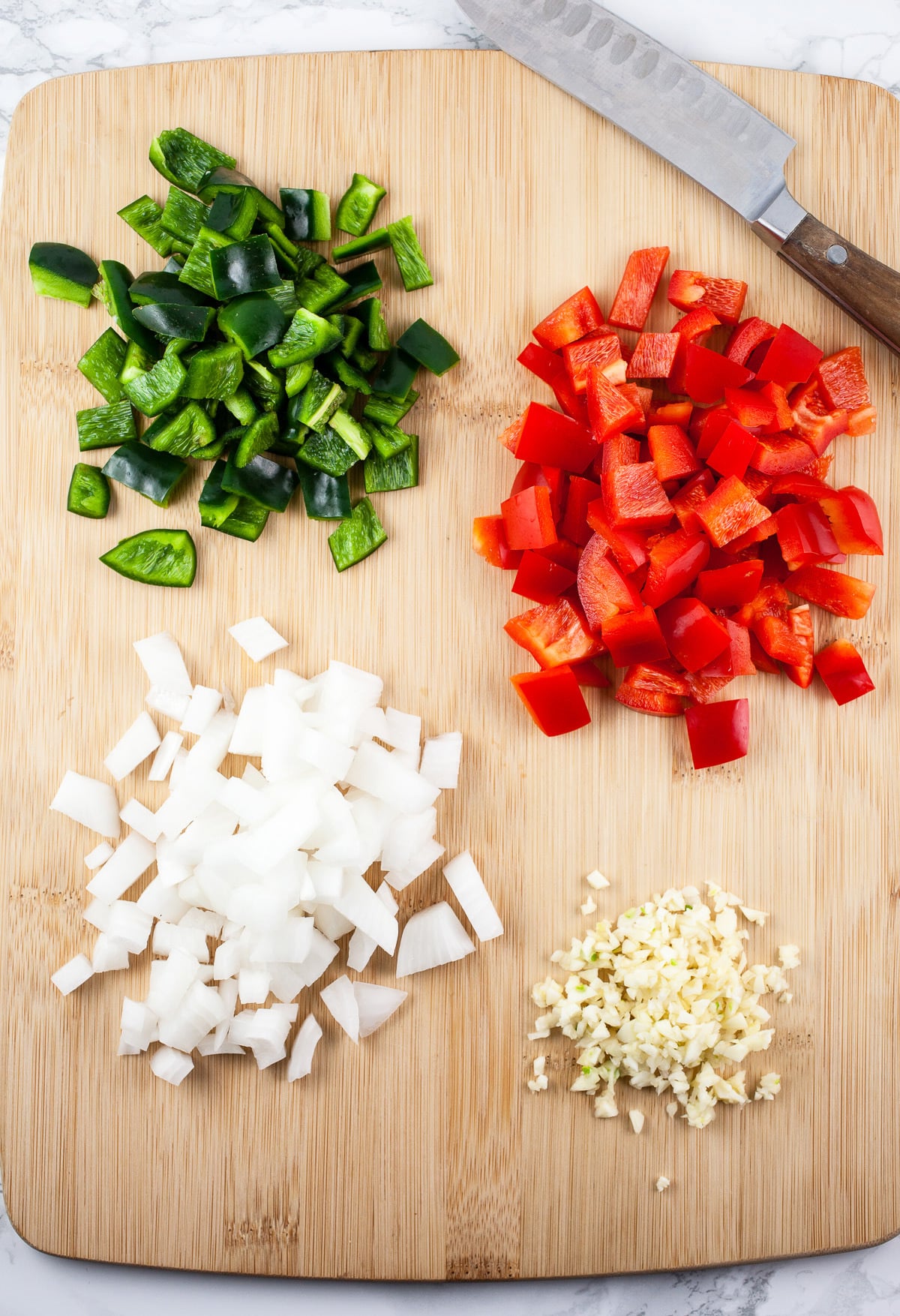 Minced garlic, onions, red bell peppers, and poblano peppers on wooden cutting board with knife.