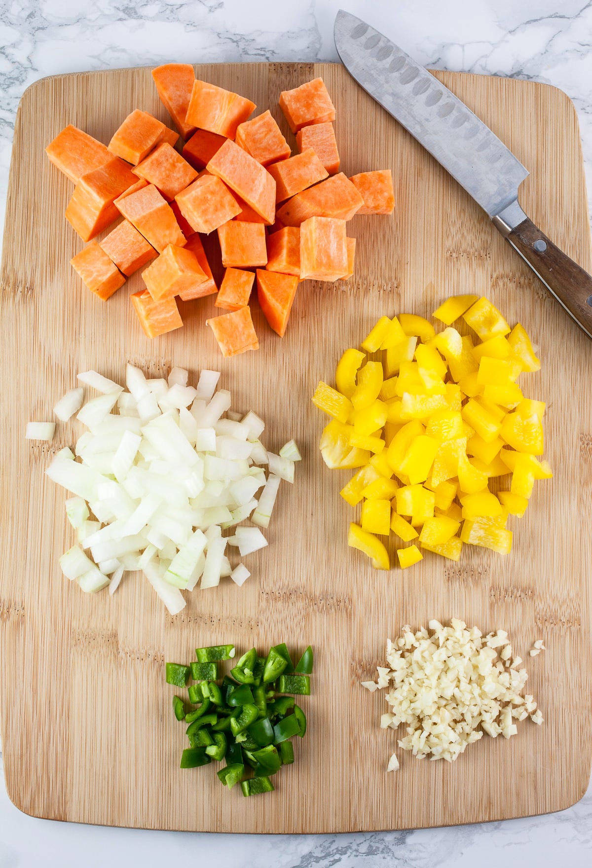 Minced garlic, jalapeno, onion, yellow bell pepper, and chopped sweet potatoes on wooden cutting board with knife.
