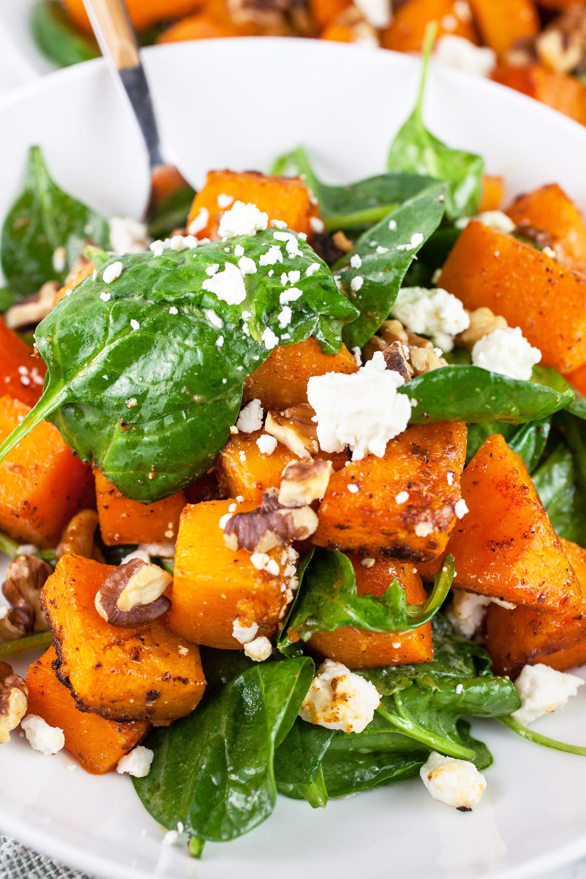 Butternut squash salad with greens, walnuts, and feta in white bowl with fork.