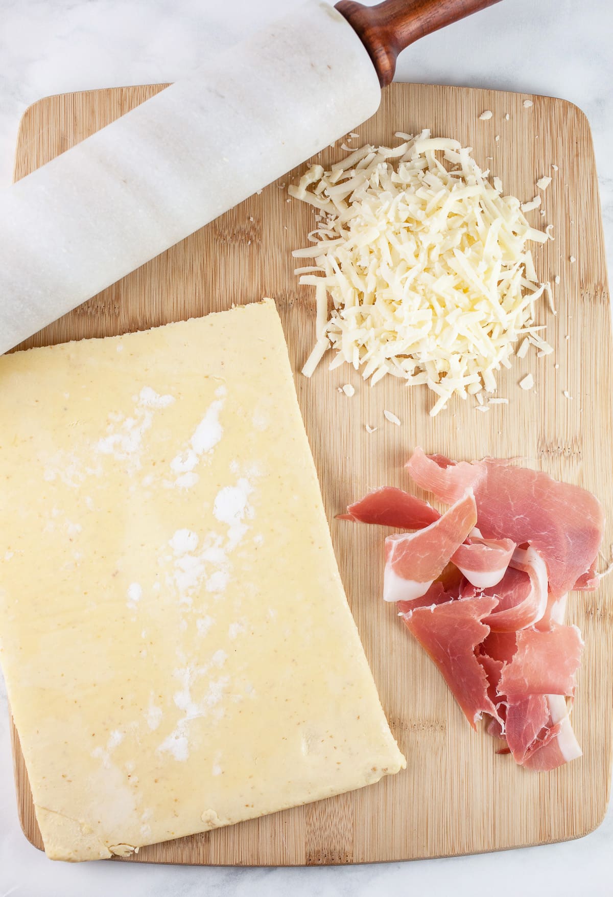 Gluten free puff pastry sheet, prosciutto, shredded gruyere cheese, and rolling pin on wooden cutting board.