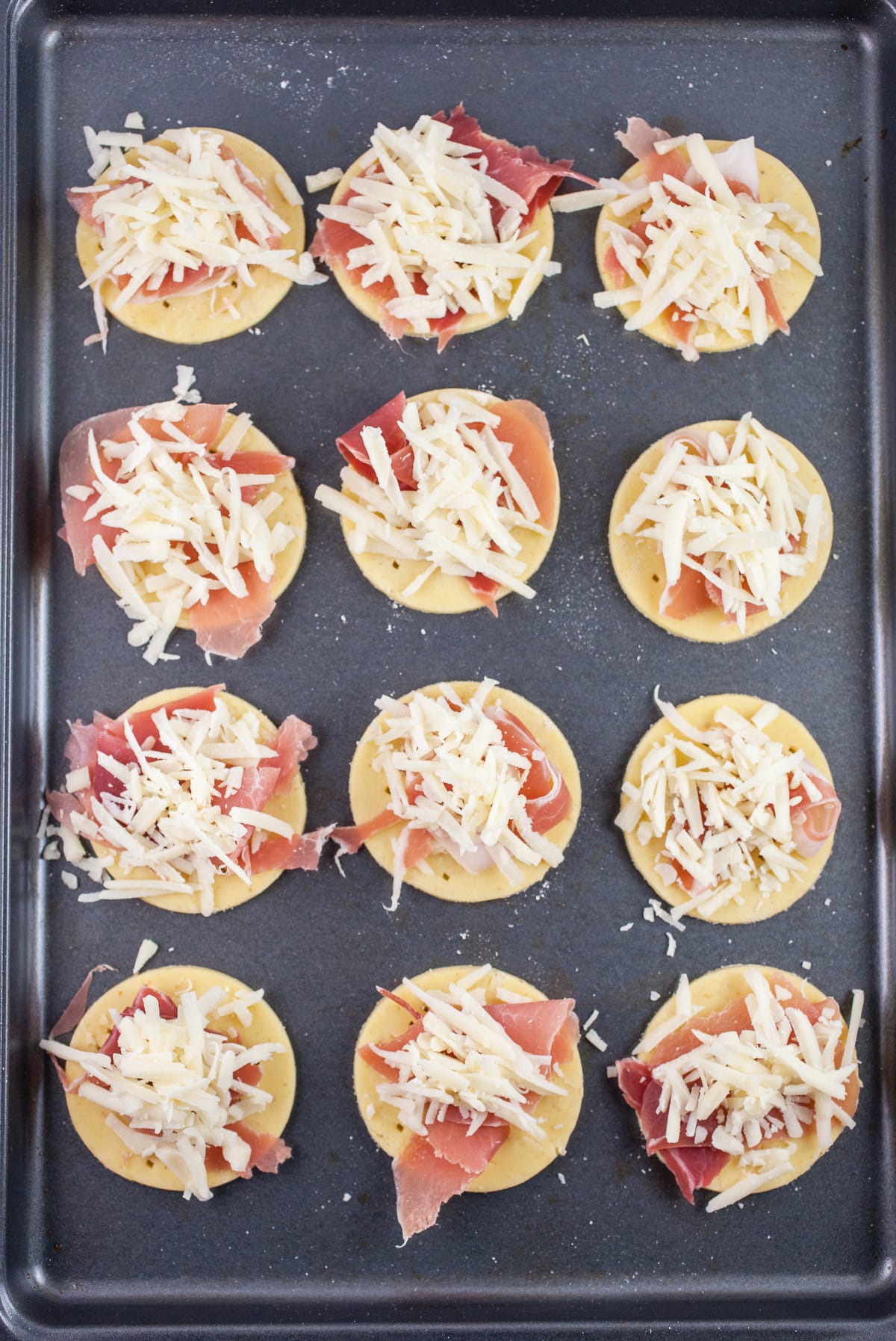 Puff pastry rounds with prosciutto and shredded gruyere cheese on baking sheet.
