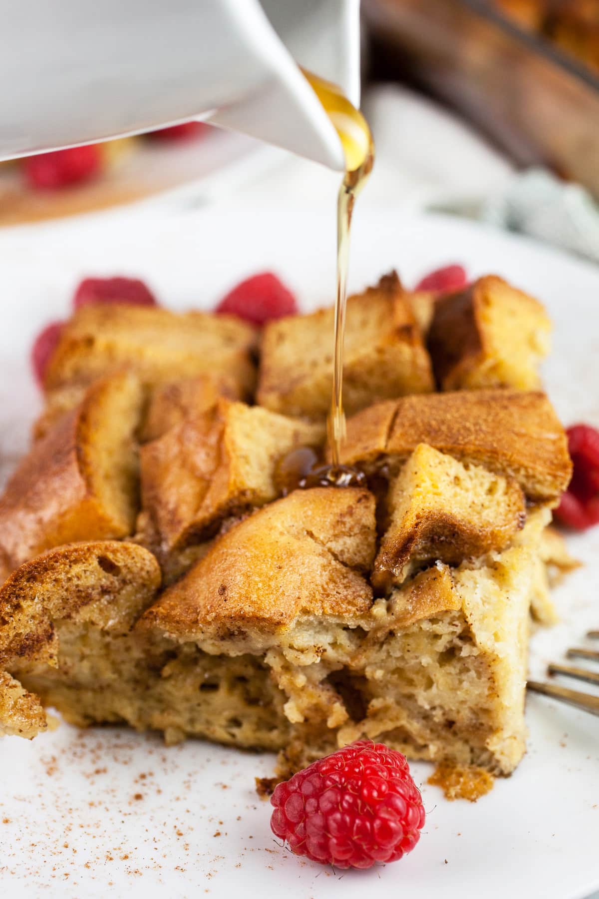 Maple syrup poured onto French toast casserole on white plate with raspberries.