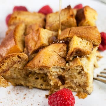 Maple syrup drizzled on piece of French toast casserole on white plate with raspberries.