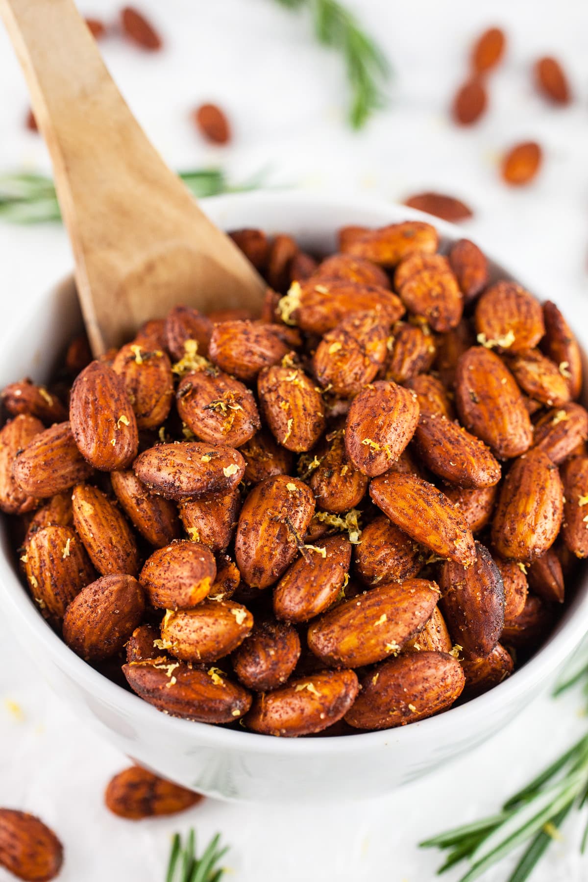 Roasted almonds in white bowl with wooden spoon next to fresh rosemary.