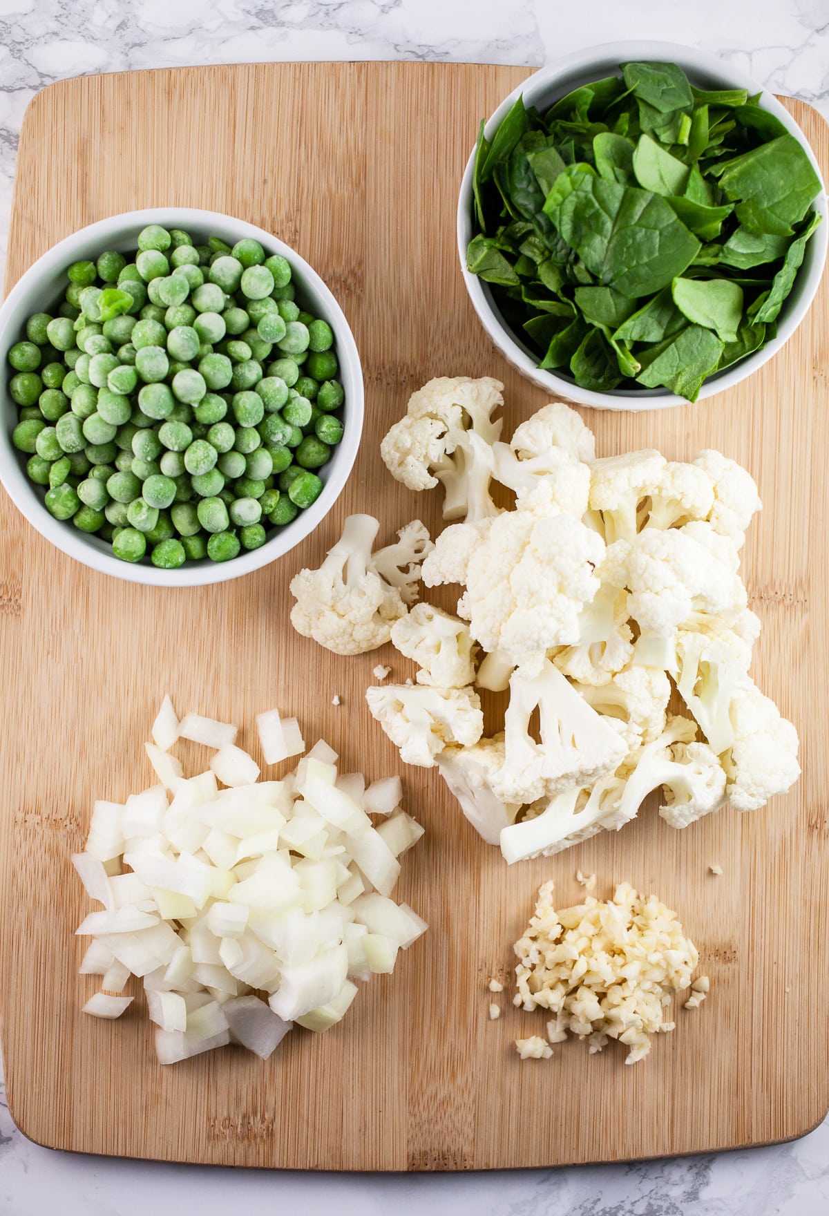 Minced garlic, onions, cauliflower florets, frozen peas, and chopped spinach on wooden cutting board.