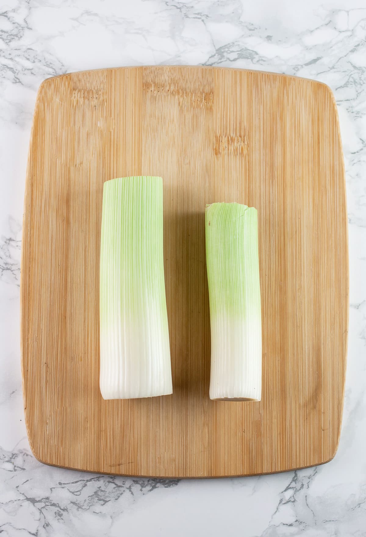 Leeks with tops and bottoms removed on wooden cutting board.