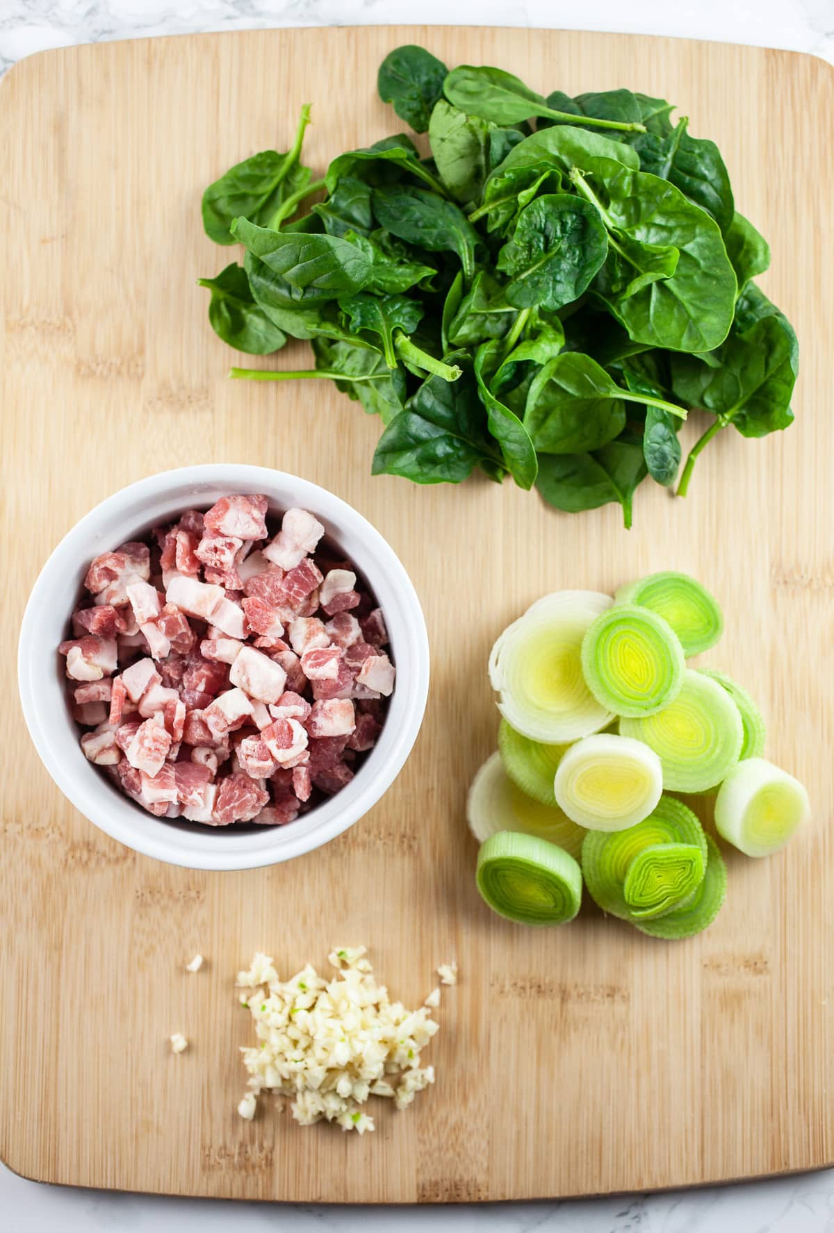 Minced garlic, sliced leeks, uncooked pancetta, and spinach on wooden cutting board.
