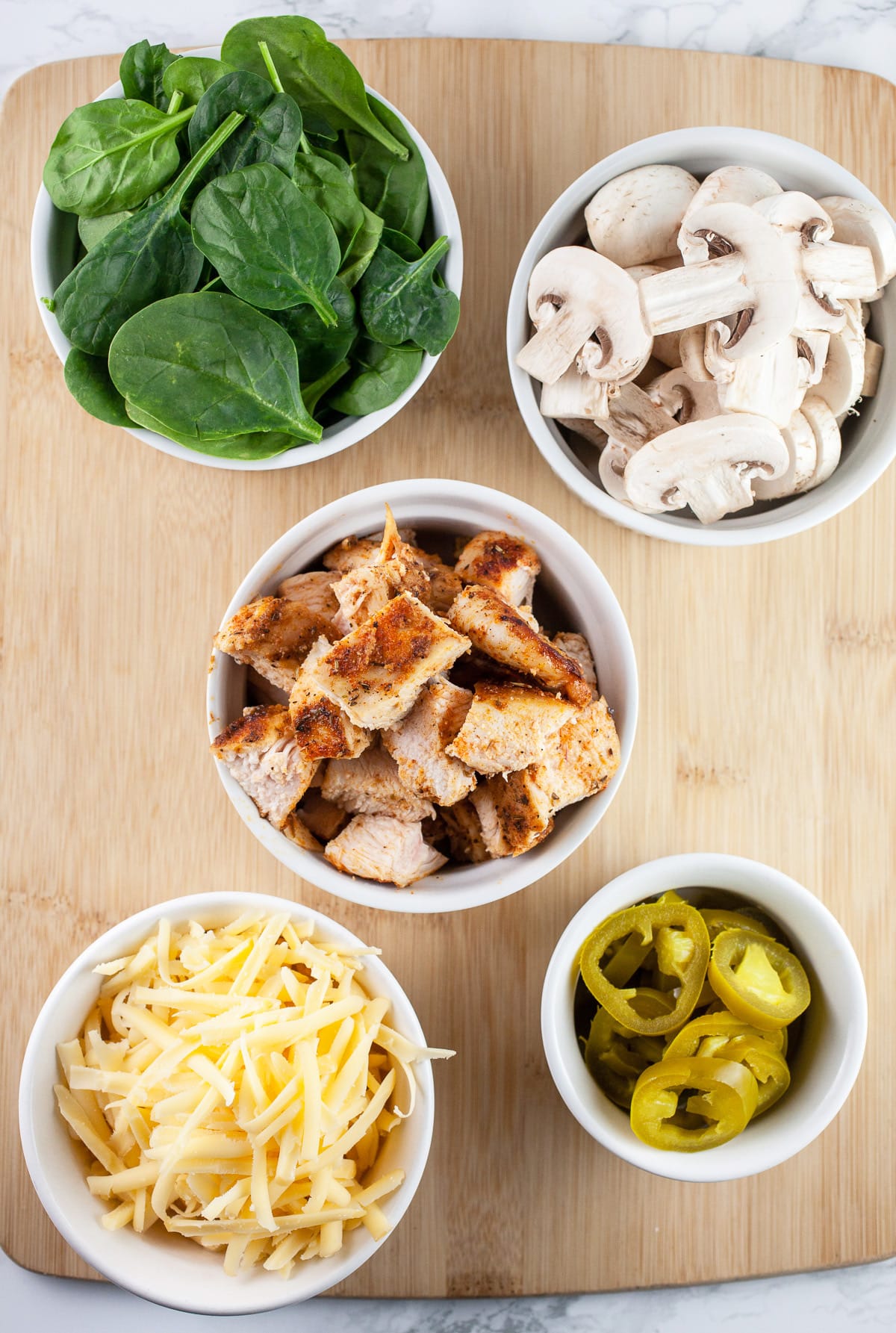 Shredded Gouda cheese, pickled jalapenos, diced chicken, spinach, and sliced mushrooms on wooden cutting board.