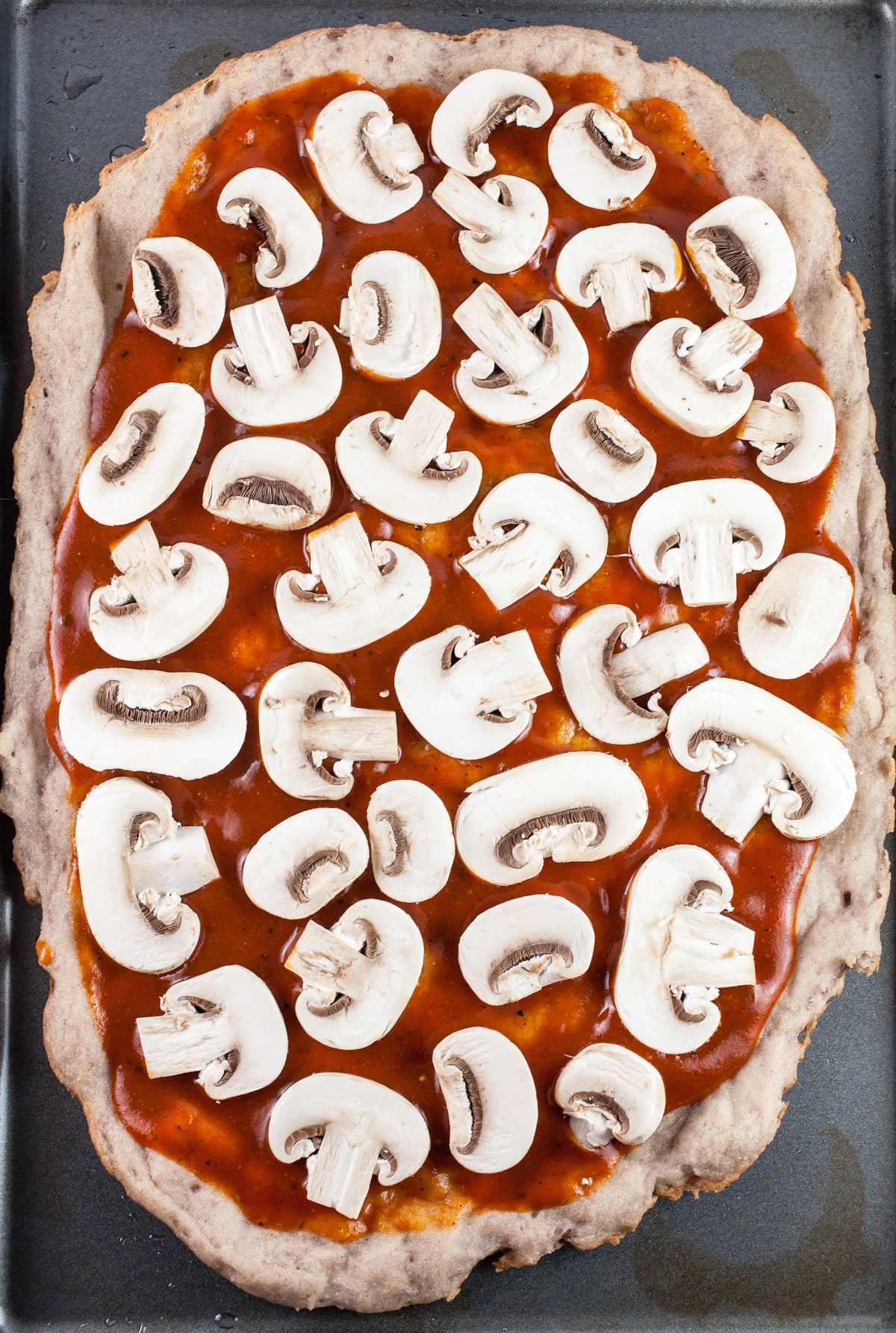 Pizza crust with BBQ sauce and sliced mushrooms on baking sheet.
