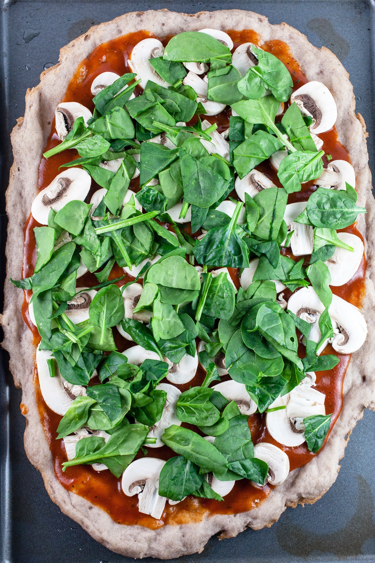 Pizza crust with BBQ sauce, sliced mushrooms, and spinach on baking sheet.