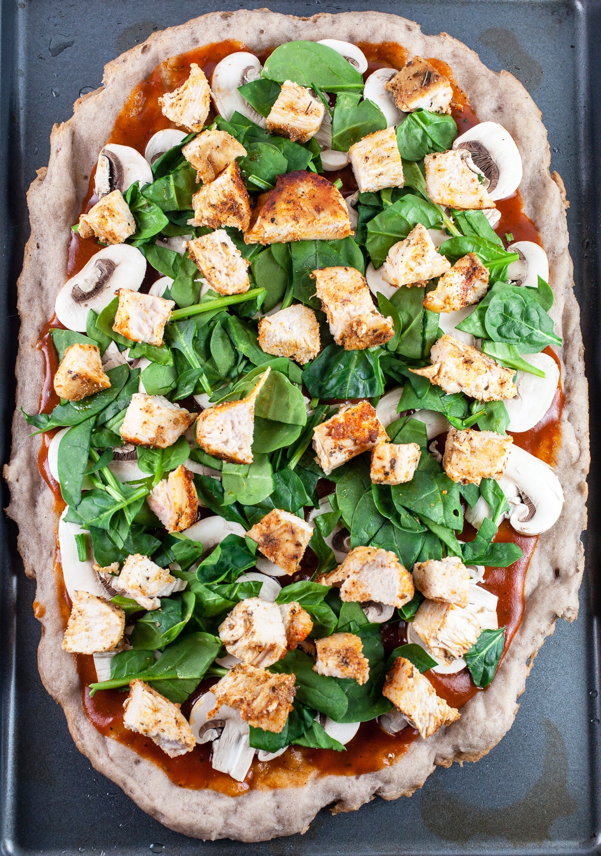 Pizza crust with BBQ sauce, mushrooms, spinach, and diced chicken on baking sheet.
