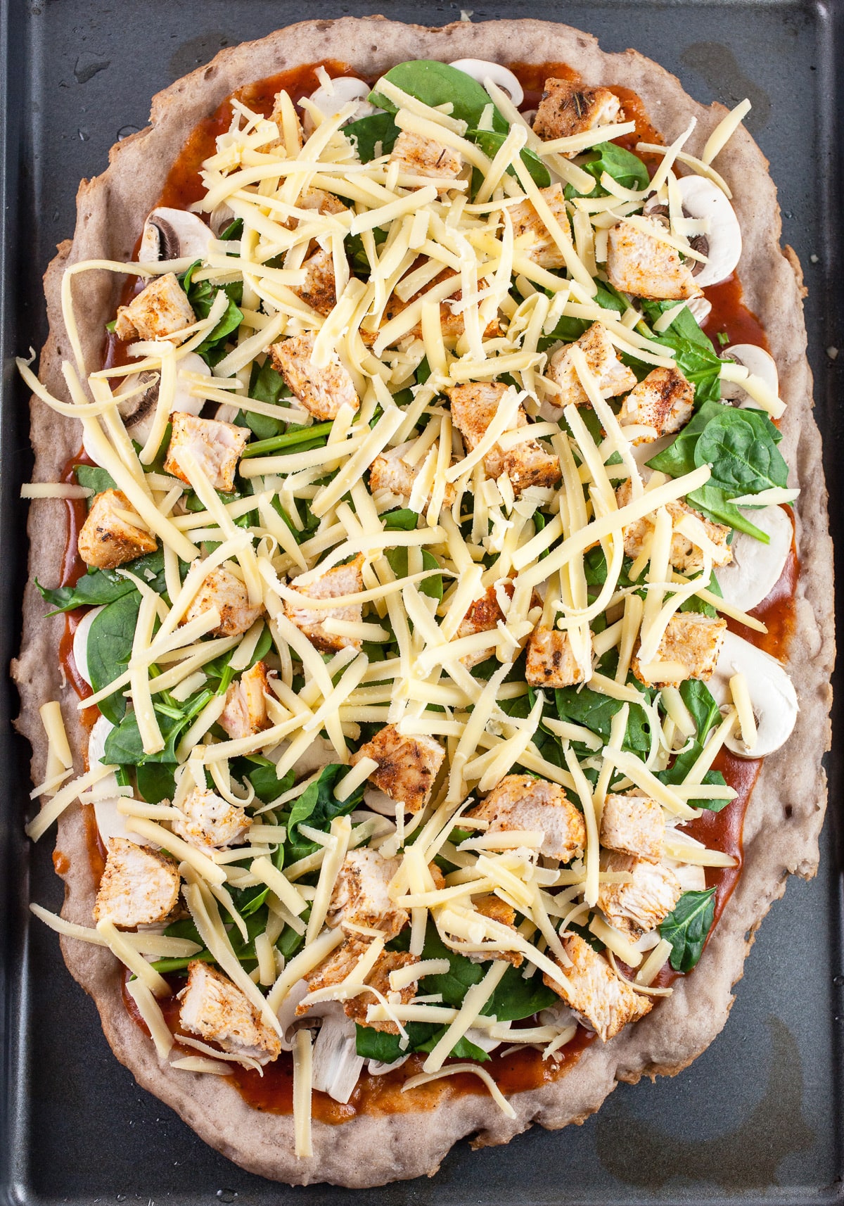 Pizza crust with BBQ sauce, mushrooms, spinach, and shredded cheese on baking sheet.