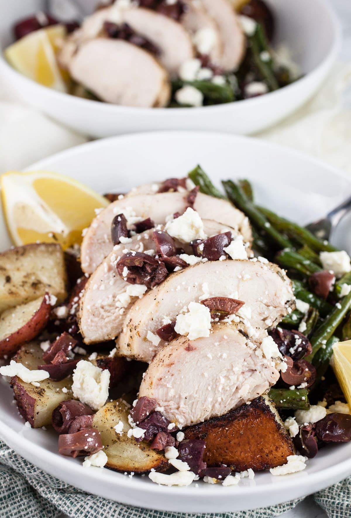 Sheet pan Greek chicken with potatoes, green beans, Kalamata olives, feta, and lemon wedges in white bowls.