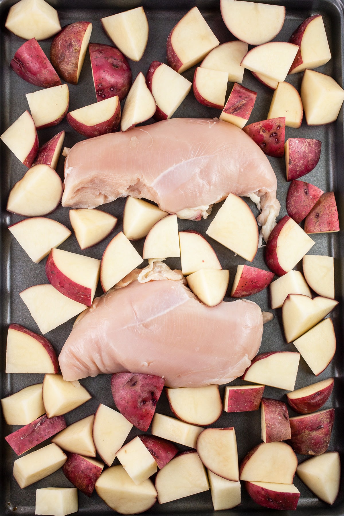 Uncooked chicken breasts and diced red potatoes on baking sheet.