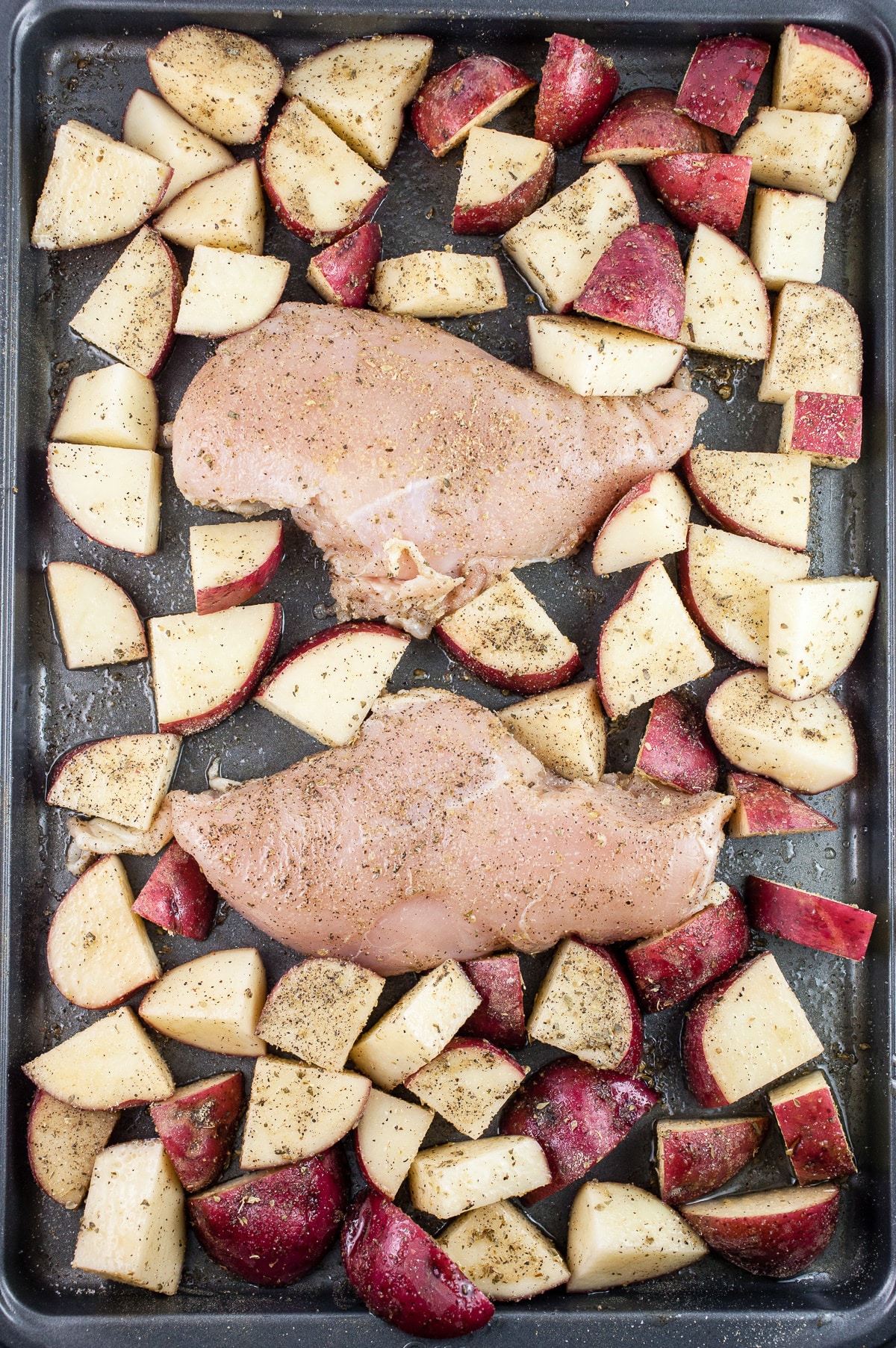 Uncooked chicken and diced red potatoes with olive oil and spices on baking sheet.