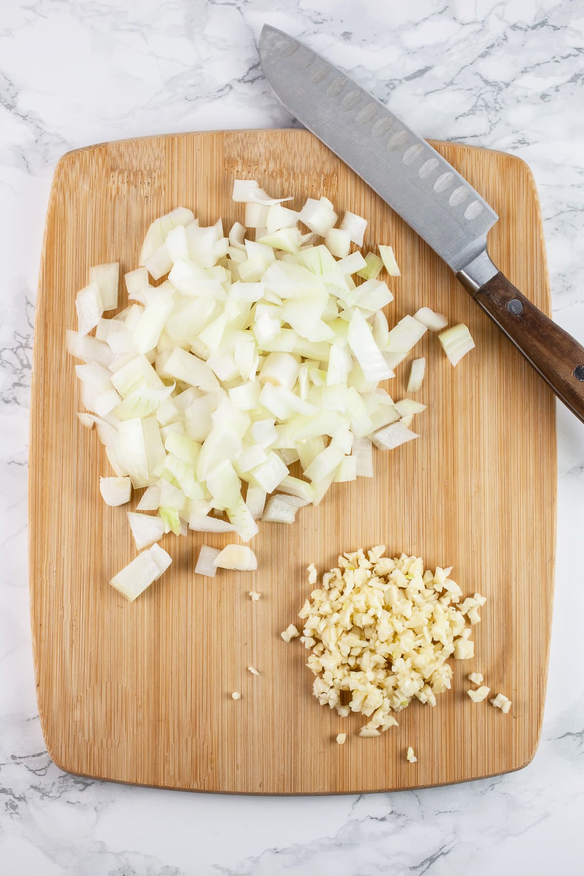 Minced garlic and onions on wooden cutting board with knife.