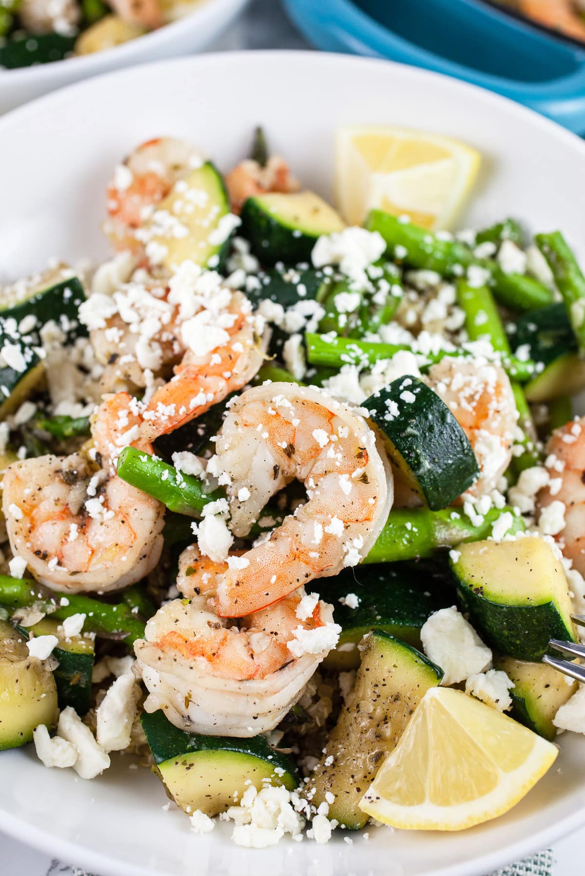Skillet shrimp with asparagus, zucchini, feta cheese, and lemon wedges in white bowls.