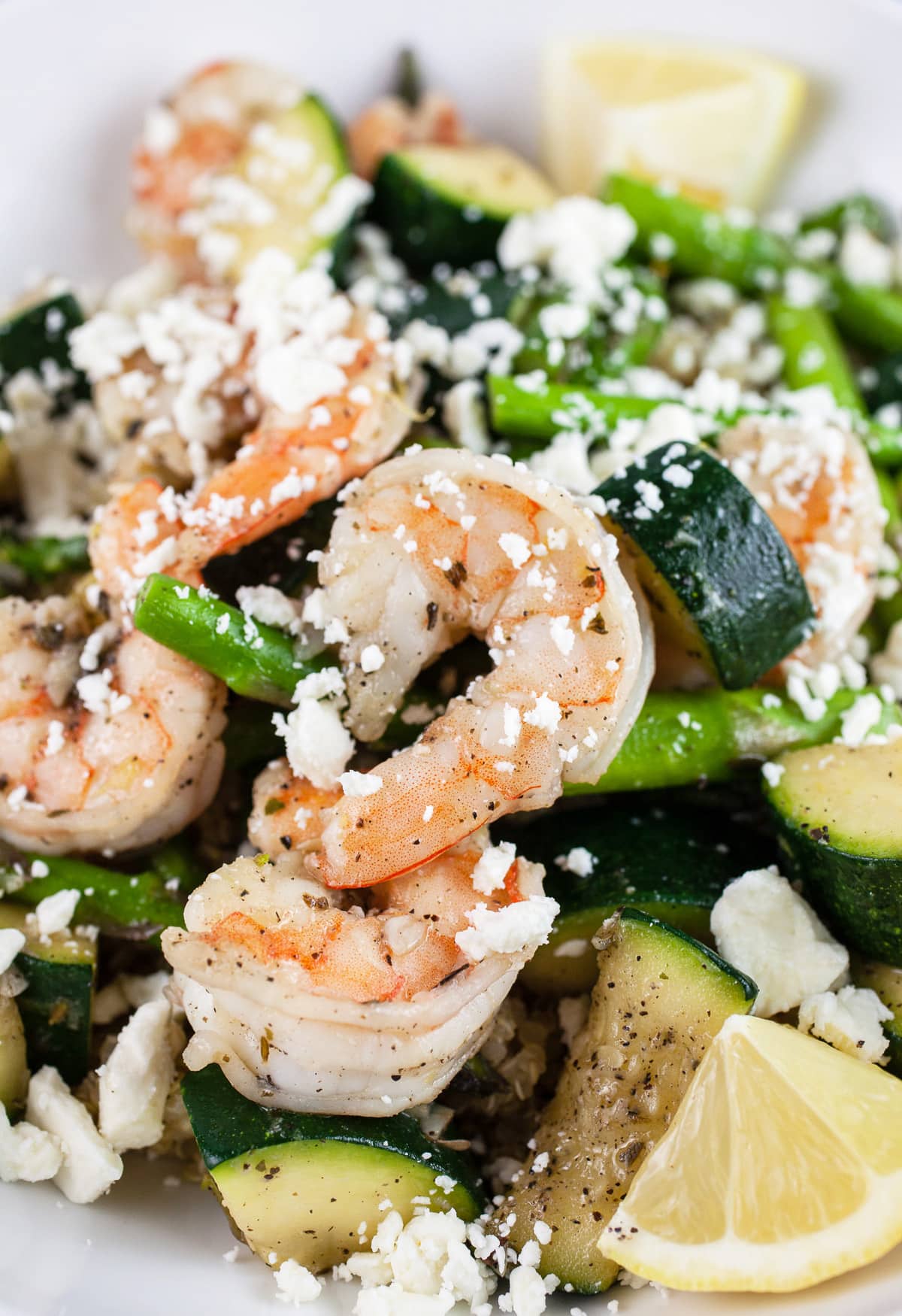 Skillet shrimp with asparagus, zucchini, feta cheese, and lemon wedges in white bowl.