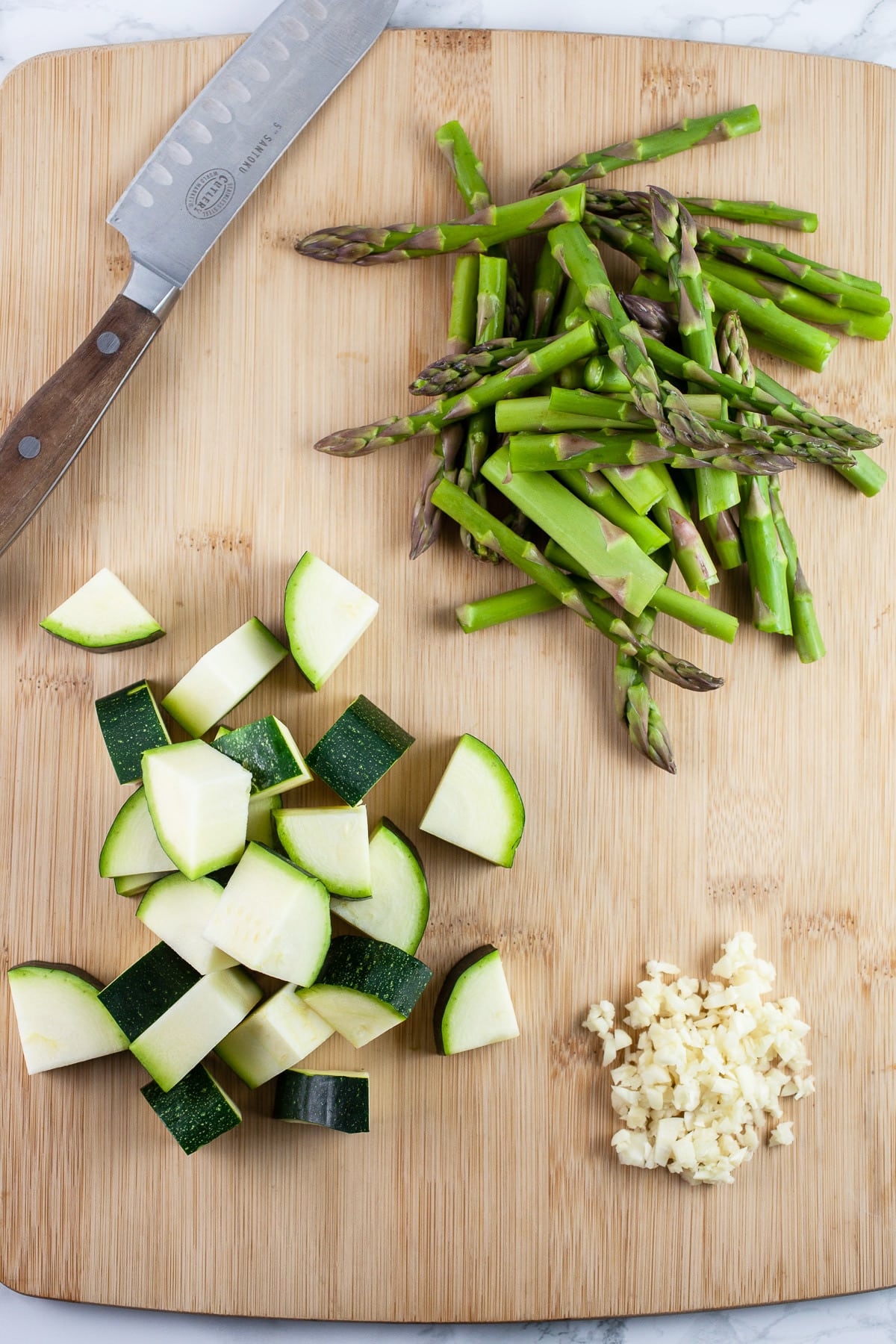 Minced garlic, diced zucchini, and chopped asparagus on wooden cutting board with knife.