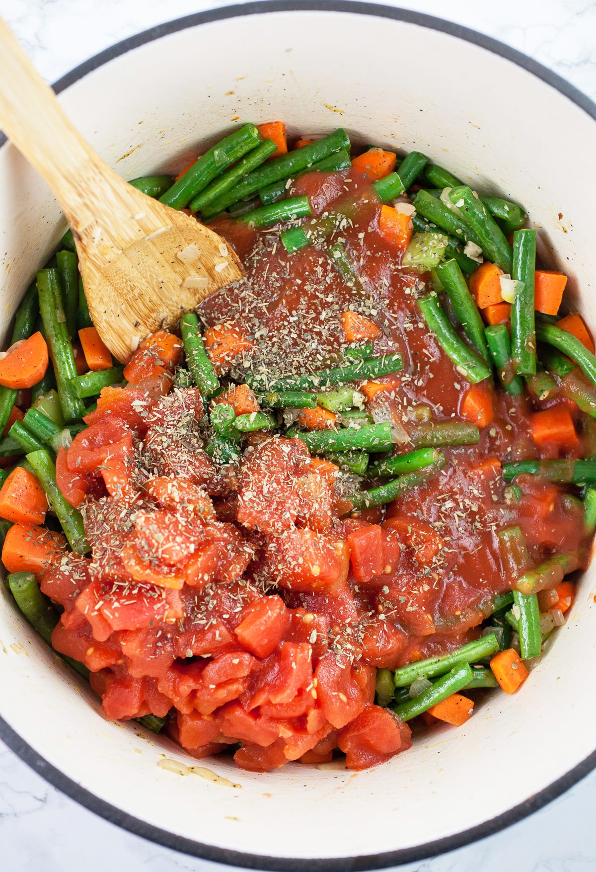 Diced tomatoes, tomato sauce, and Italian seasoning added to vegetables in Dutch oven with wooden spoon.