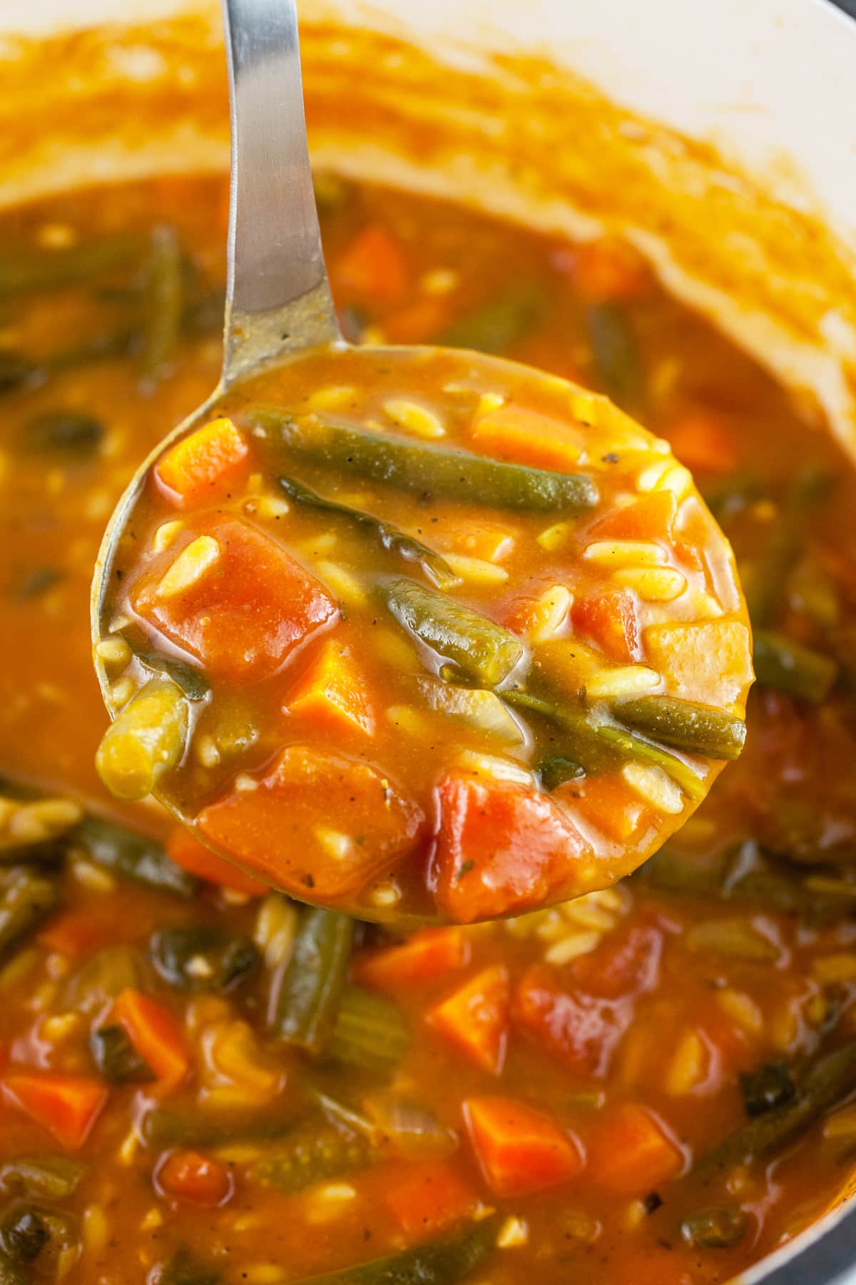 Scoop of orzo vegetable soup lifted from Dutch oven on metal ladle.