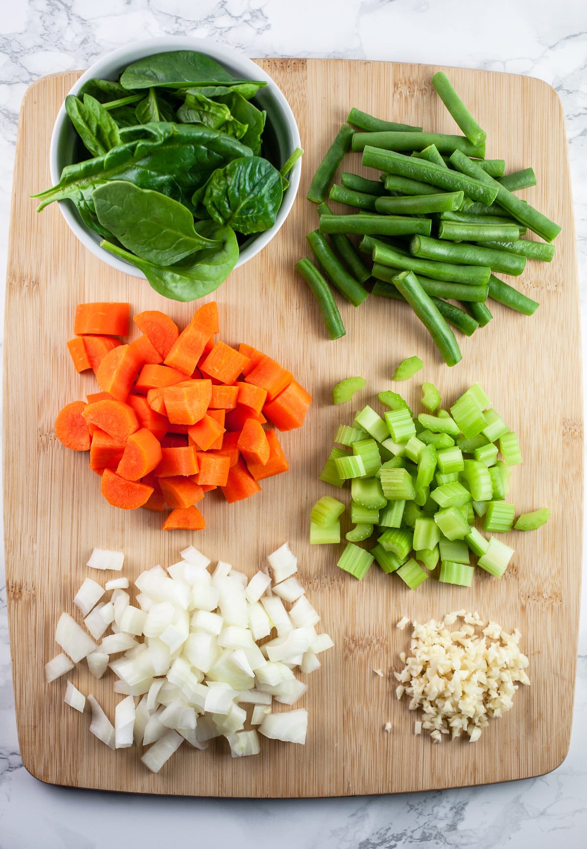 Minced garlic, onions, celery, carrots, green beans, and spinach on wooden cutting board.