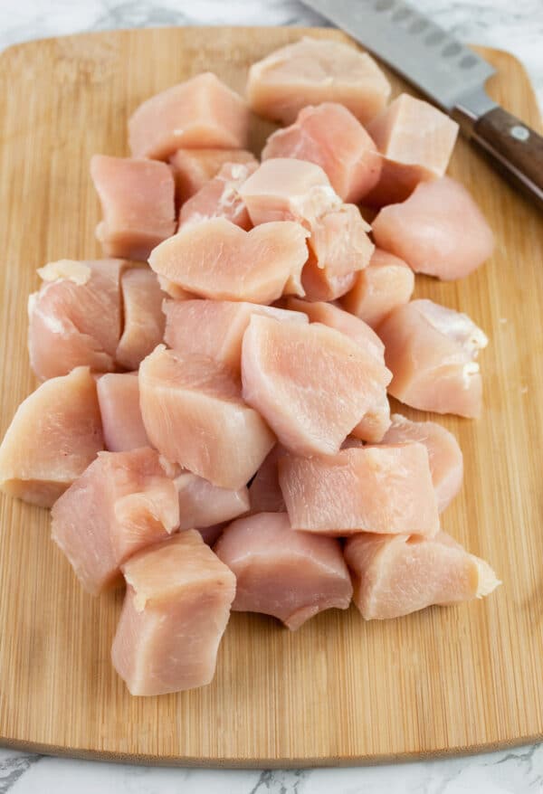 Raw chicken breasts cut into chunks on wooden cutting board with knife.
