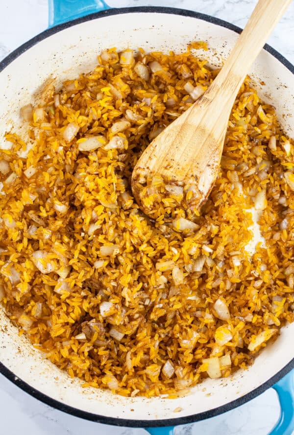 Garlic, onions, spices, and uncooked orzo pasta sautéed in skillet with wooden spoon.