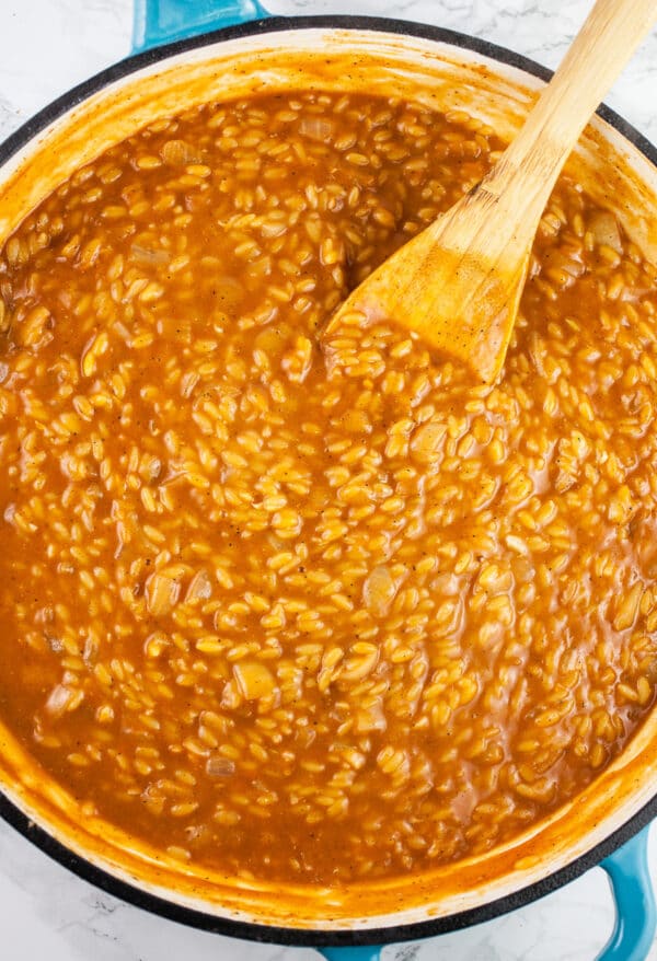 Orzo pasta cooked in tomato broth in skillet with wooden spoon.