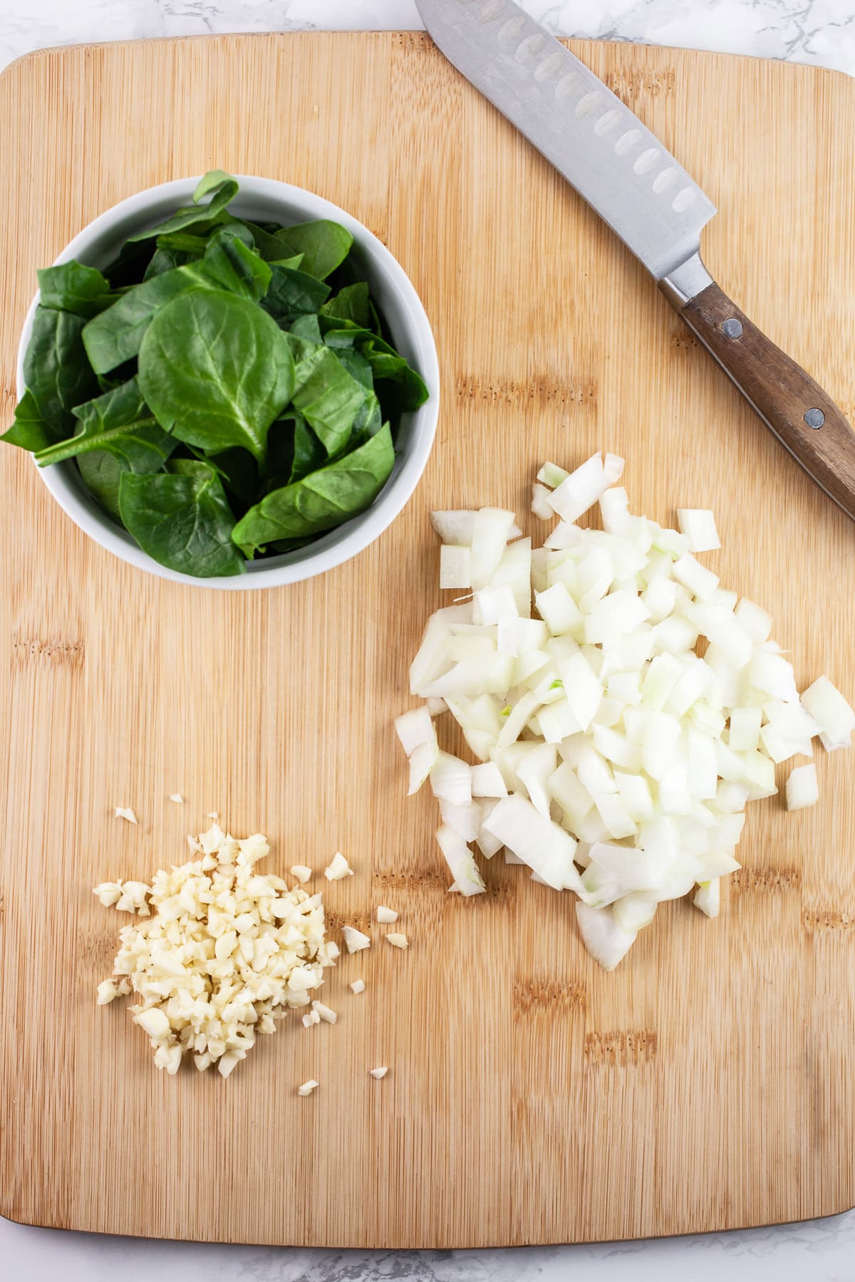 Minced garlic, onions, and spinach on wooden cutting board with knife.
